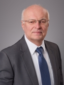 Headshot of male with white hair and eyeglasses wearing a grey suit, white shirt, and blue patterned tie.