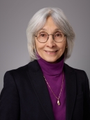 Headshot of female with short grey hair wearing a purple turtleneck, black jacket and a necklace.