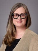 Woman with long brown hair with eyeglasses wearing a blouse under a light brown sweater.