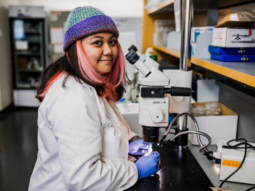 Young woman with pink hair and a purple and green beanie works in a lab wearing a white lab coat.