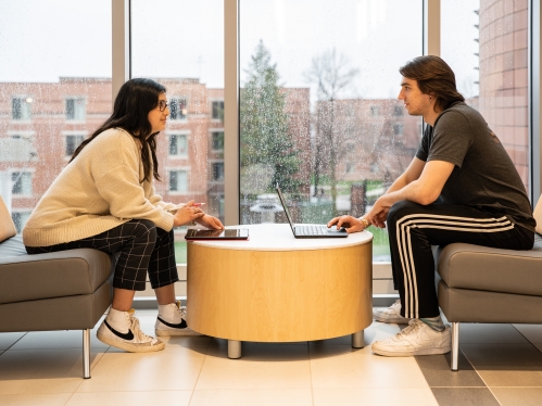 Male and female students face each other across a low round table. Each have a laptop.