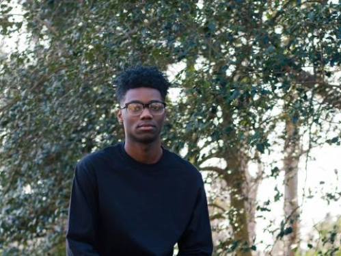 African American student with glasses, wearing a black shirt and gray pants, sits on a rock outdoors.
