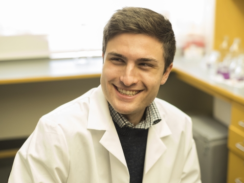 Young white male smiling with brown hair posing in a chemistry lab wearing a white lab coat.