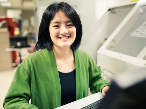 Asian young woman with black hair, smiling in a lab, wearing a bright green sweater over a black shirt.