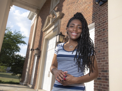 Young black woman posing in front of a brick building. She is wearing a blue and white striped shirt, smiling with her hair in long braids.