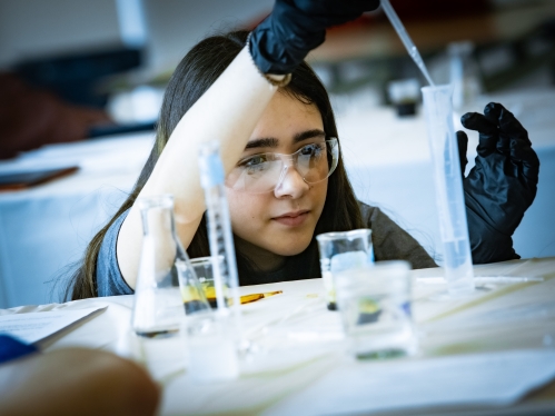 Female student wearing safety goggles and black gloves uses a pipette and chemistry tube.