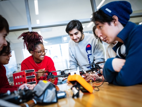 A group of six students, male and female gathered around a wooden table working on a drone.