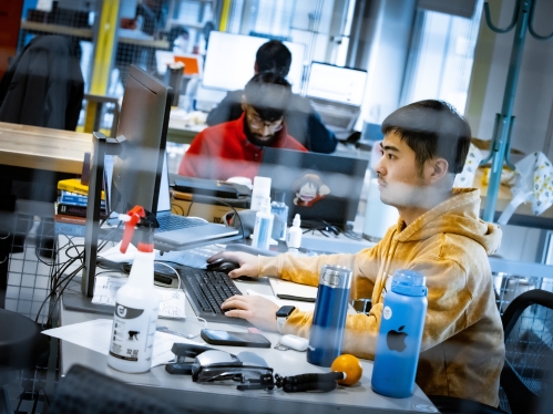 Male student in yellow sweatshirt working at a computer with other students in background.