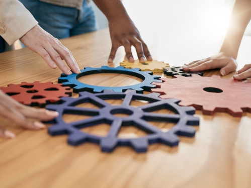 Painted gear puzzle pieces on a wooden desk; hands touching the pieces.