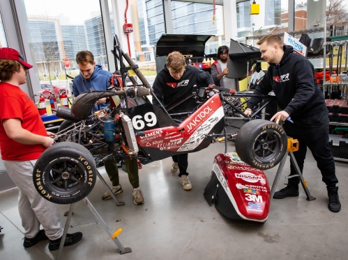 Four male students work on a formula race car in an open glass-walled garage.