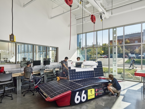 Studios work in garage space with a wall of windows. A solar car is in the center of the space.