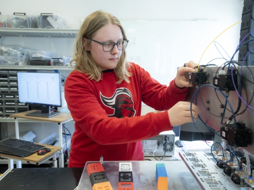College student wearing a red long-sleeved shirt, blond hair and glasses works on a 3D printing machine.