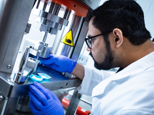 Male student with black hair, beard and glasses, wearing blue gloves and lab coat examines pills in a testing facility.