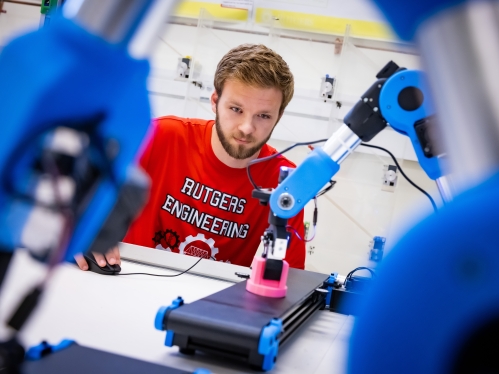 Male student wearing a red T-shirt sitting at a table behind robotic arms and conveyor belt conducting research.