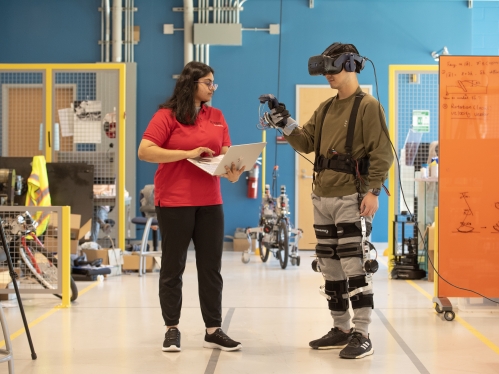 A male and a female student work in a lab. She is wearing a red shirt and black pants holding a laptop. He is wearing a VR headset with devices attached to his legs and hands.