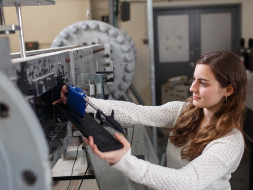 Female student with long brown hair wearing a white shirt puts a device inside a wind tunnel.