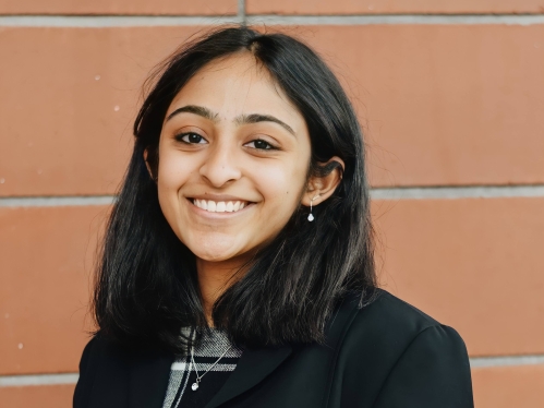 Young woman poses in front of a brick wall. She has black hair and is wearing a black jacket.