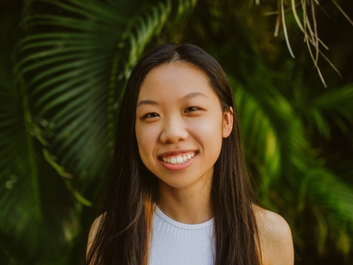 Female college student poses outside in front pine trees. She has long dark hair and is wearing a white sleeveless top.