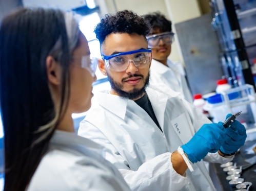 Two male students and one female student working in lab wearing safety goggles and a white lab coats.