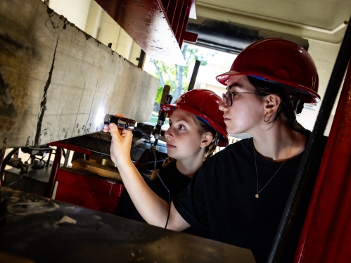 Two female students in red hard hats use a flashlight to exam concrete slab in a structural lab.