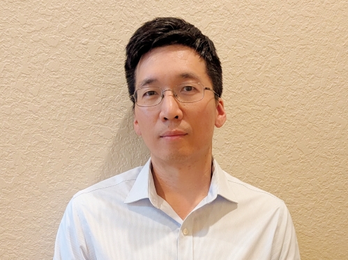 Headshot of man with black hair and glasses wearing an open collared white shirt.