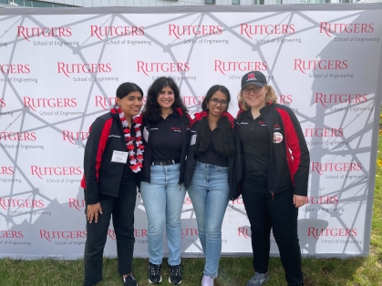 Four female students wearing Rutgers black and red jackets stand in front of a white backdrop with Rutgers Engineering logo repeated.