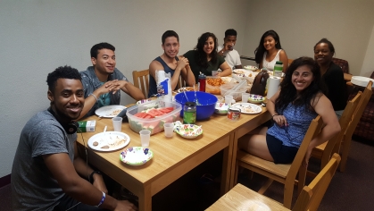 Eight students, male and female, sit at a table where they are having lunch. All are looking at the camera and smiling.