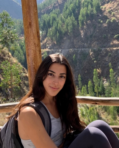 Young woman with long dark hair sitting with a back pack with a view of trees and mountains behind her.