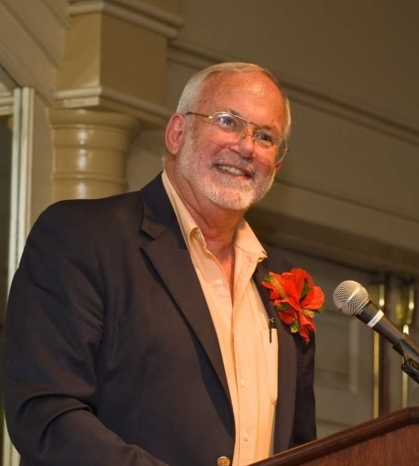 Older white man with silver hair with glasses. Speaking from a podium wearing a dark suit jacket, yellow shirt and orange lapel flower.