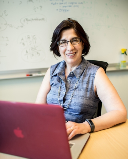 Woman wearing a sleeveless blue plaid collared top smiles at the camera as she sits at a table with a laptop open.