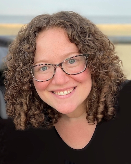 Headshot of smiling young woman with curly brown hair, blue eyes and glasses.