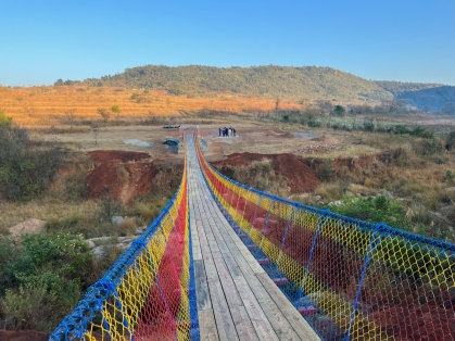 View of a footbridge with colorful roping, looking across the bridge with mountains and blue sky in the background.