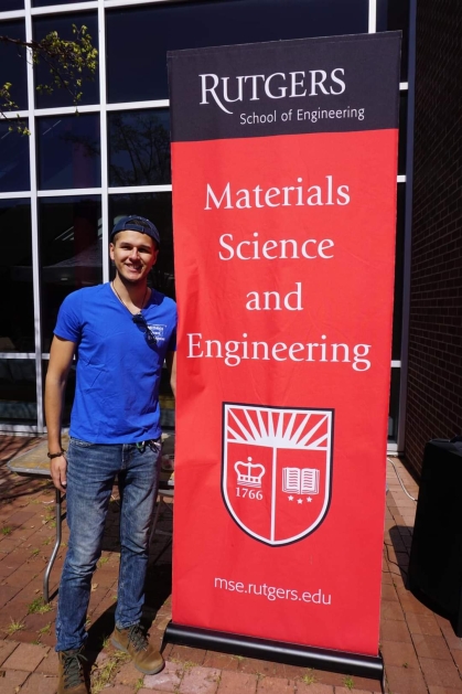 Male college student wearing a blue polo shirt, baseball cap and blue jeans stands next to a vertical banner that says Rutgers Materials Science and Engineering.