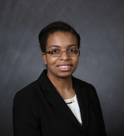 Head shot of young black college student. She is wearing a black suit jacket with a white top underneath. Her hair is short and she has glasses.