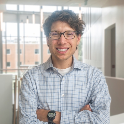 Head shot of white male student. His arms are crossed, he's smiling with glasses, brown wavy hair and wearing an open neck blue collared shirt.