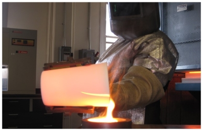 Demonstration of molten glass being poured from a beaker by a person in hood and protective clothing.