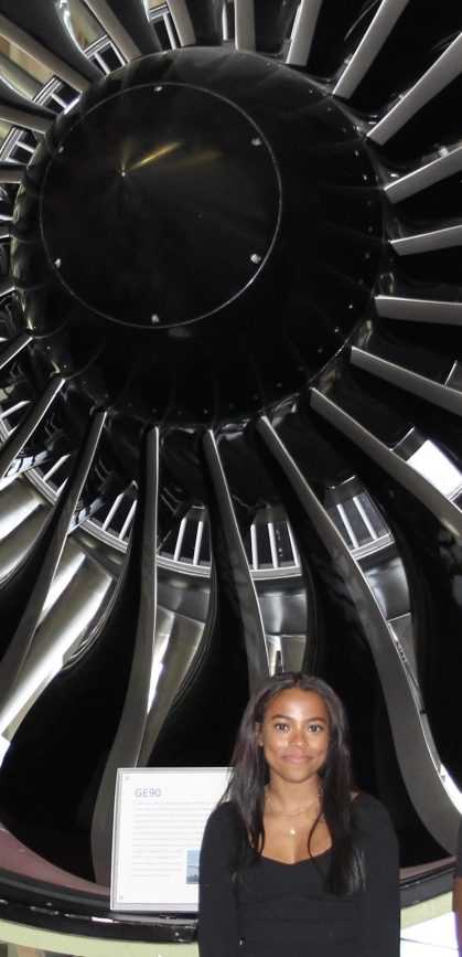 Woman with black hair and black shirt stands in front of a airplane engine.