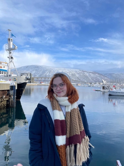 Young woman with red hair wearing a heavy coat and brown striped scarf stands near a harbor in Norway with fishing boats and mountains in the background.