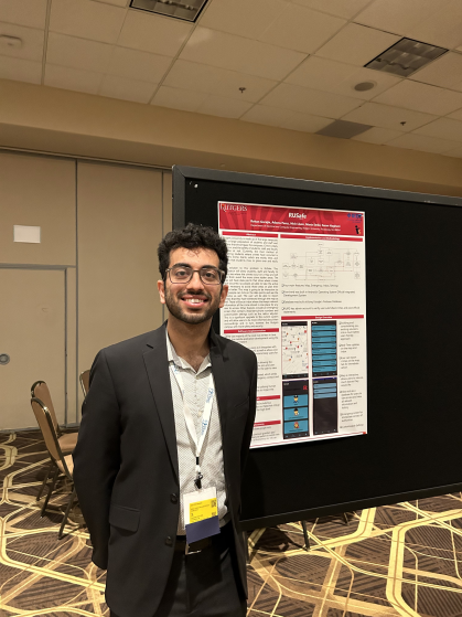 Male student with eyeglasses and a beard wearing a dark grey suit and white button down shirt standing in front of his research poster. 