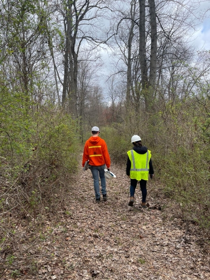 Two people are pictured from behind as they walk along a path in the woods. One is wearing a red jacket and white hat. The person on the right has on a yellow reflective vest and a white hardhat.