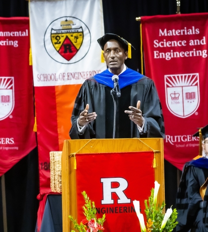 Black male wearing graduation robes, stands behind a podium with banners behind him.