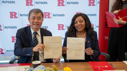 Man and woman hold signed certificates while seated at a conference table in front of a Rutgers backdrop.