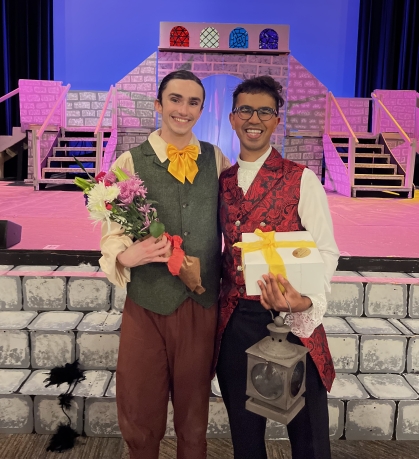 Two young men wearing period costumes pose on a stage holding flowers and a gift. The backdrop is a play setting.