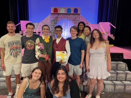 Eight young men and women gather around two male actors is period costumes, standing on stage with scenery background.