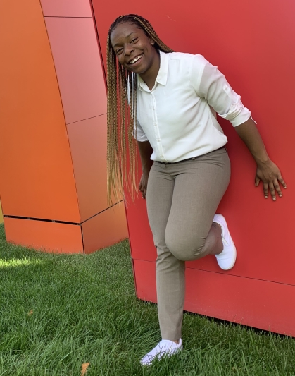 Female student with brown skin and long braids wearing a white blouse with khaki pants, leans against a red wall with one leg up.