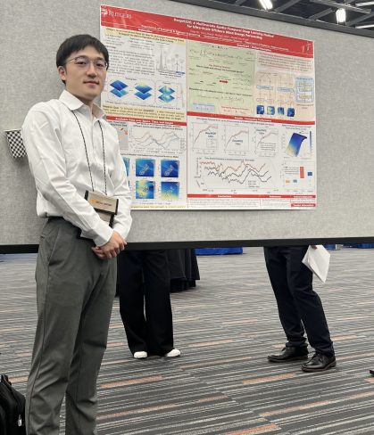 Male student with black hair wearing a white shirt and dark slacks, stands next to a research poster.