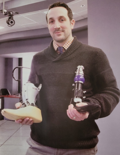 Male wearing a brown sweater with a button down shirt, holding two prosthetic devices.