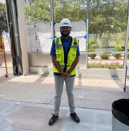 Male college student is on a construction site wearing a hard hat, yellow vest, and light colored pants.