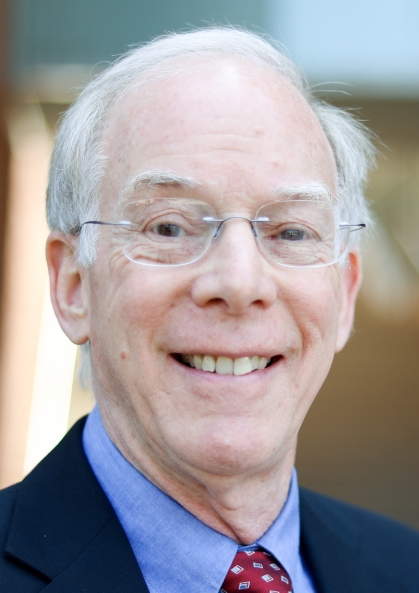 Headshot of male with white hair and glasses, wearing a blue suit, blue shirt and red tie.