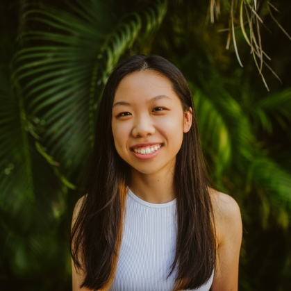 Female college student poses outside in front pine trees. She has long dark hair and is wearing a white sleeveless top.